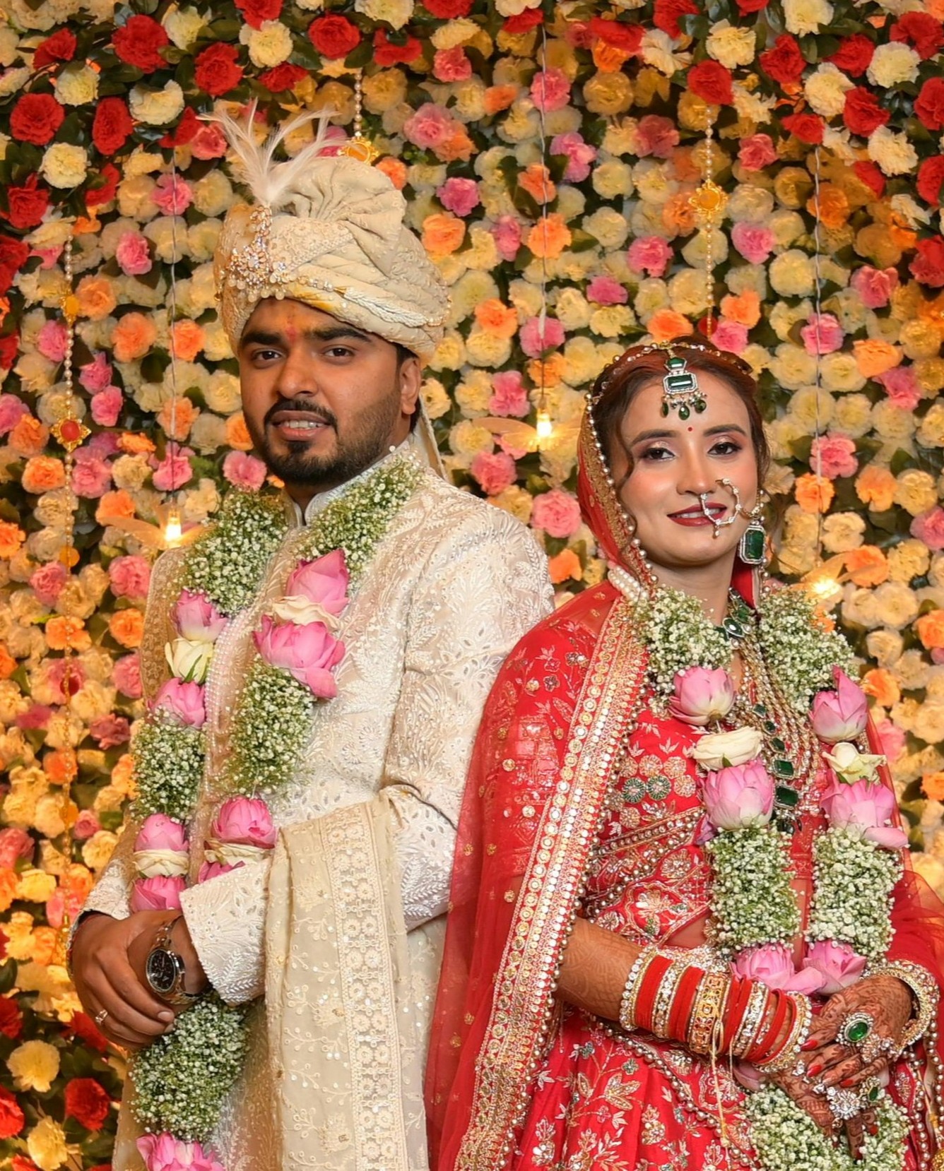 Smiling Indian bride in a red embroidered lehenga and groom in an ivory sherwani with turban, wearing rose garlands in front of a floral wall - a Hamsafar matched couple
